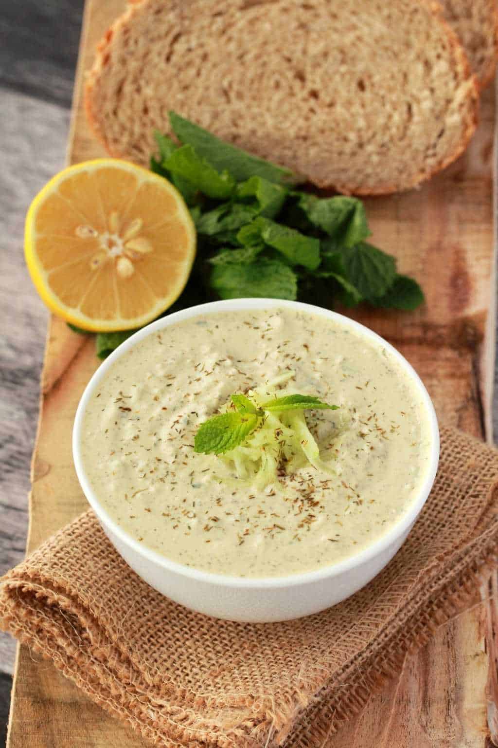 Tzatziki végétalien dans un bol blanc avec du citron frais, de la menthe et du pain tranché en arrière-plan.