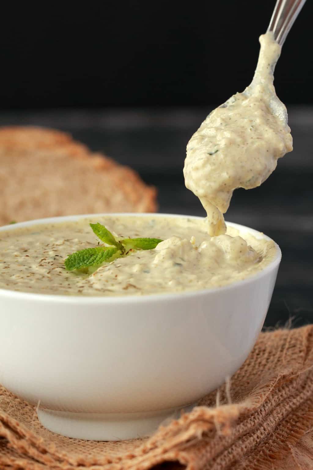 Une cuillerée de tzatziki végétalien sur un bol blanc rempli de tzatziki, du pain en tranches en arrière-plan.