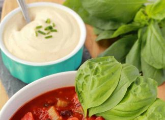 Easy Homemade Vegan Chili – Loving It Vegan Chili végétalien dans un bol blanc avec une cuillerée de crème de noix de cajou, de ciboulette hachée et de feuilles de basilic frais. Un bol de crème de noix de cajou et de basilic plus frais est à l