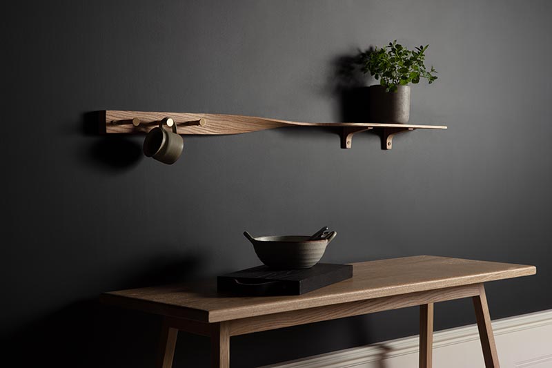 Soigneusement sculpté dans du bois de frêne ton sur ton, le Tor Twist Shelf se tord doucement au centre et s'élève au-dessus de trois patères en laiton brossé. #WoodShelving #WallShelf #ModernShelf #KitchenShelving #EntrywayShelf