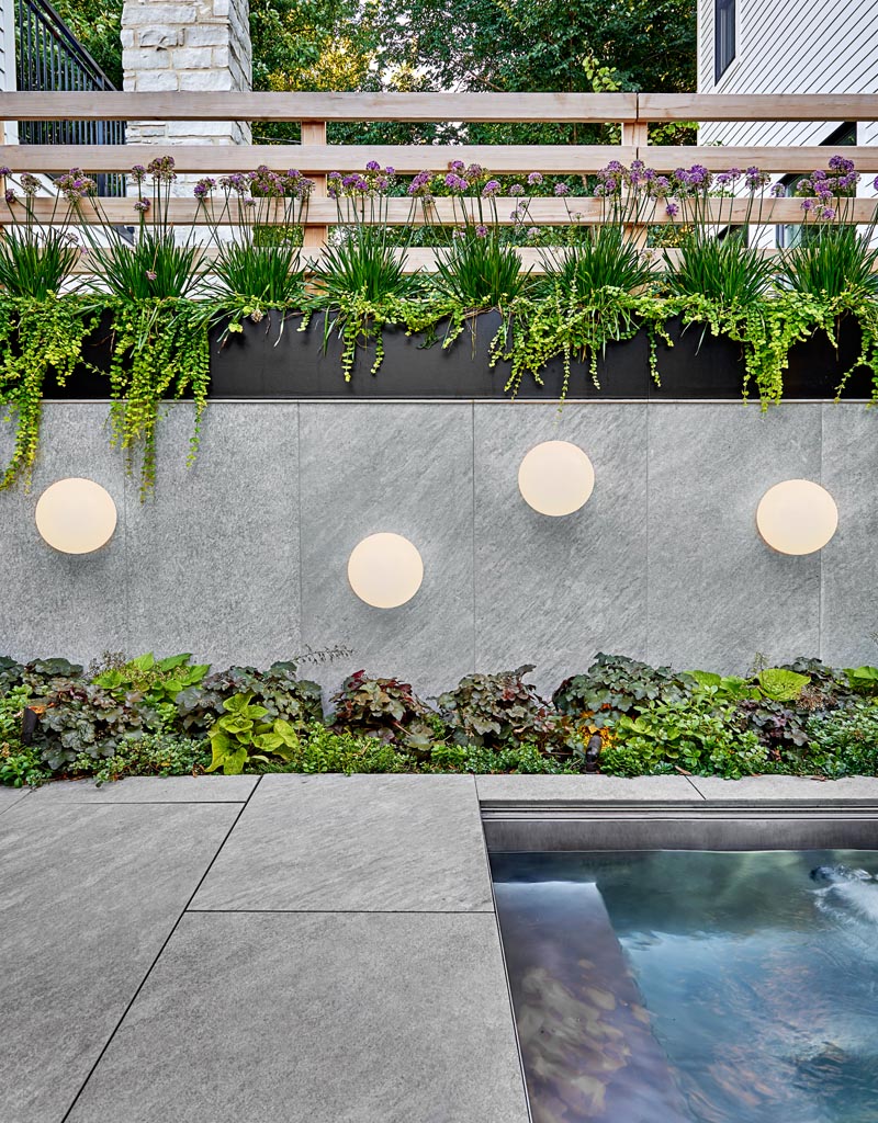 Cet espace extérieur moderne dispose d'un carreau de porcelaine aspect pierre, tandis que les plantations indigènes résistantes à la sécheresse ajoutent une touche naturelle, et les luminaires globe ajoutent une touche artistique au mur du spa conçu sur mesure. #Paysage #PlanteursAcierNoir #Éclairage extérieur # Éclairage extérieur