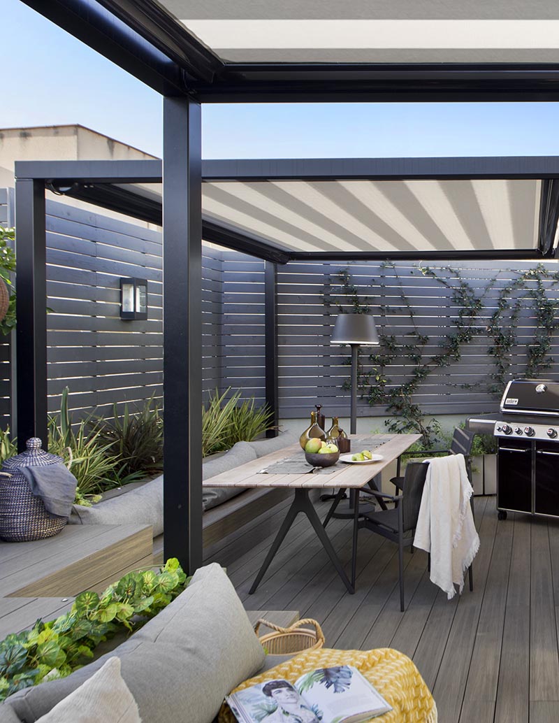 Cette terrasse extérieure moderne dispose d'un coin repas avec des banquettes assorties au salon voisin, tandis que la clôture en lattes de bois gris ajoute à la sensation moderne de l'espace. #OutdoorTerrace #OutdoorDining #OutdoorEntertaining