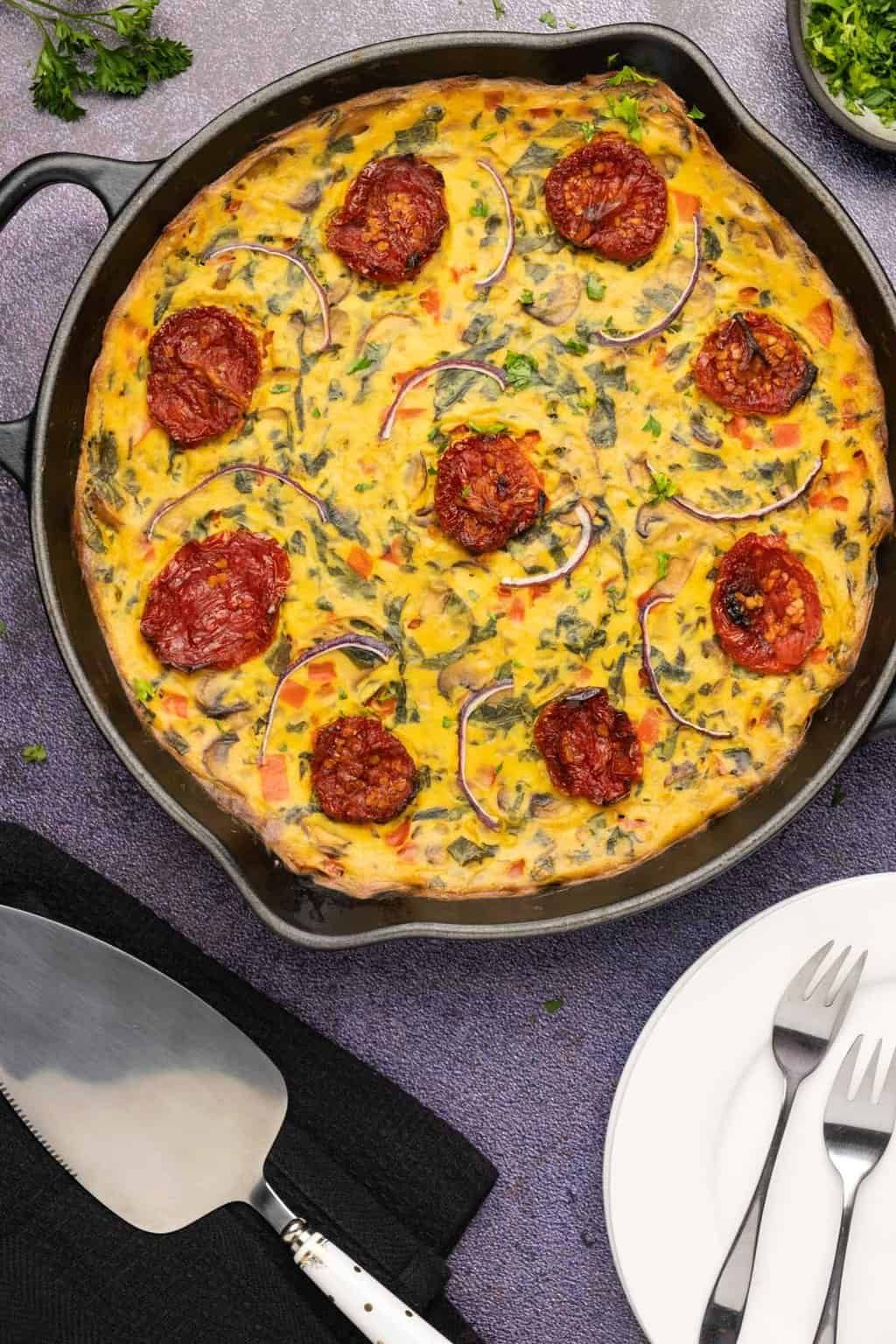 Frittata végétalienne garnie de tomates séchées et d'oignons rouges dans une poêle en fonte.