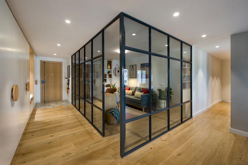 À l'intérieur de ce bureau à domicile vitré moderne se trouve un long bureau en bois qui rencontre une étagère conçue sur mesure, avec certaines des étagères bordées de bois. En face du bureau se trouve un canapé gris confortable, quelques plantes et des oreillers colorés, tandis qu'un tapis ajoute une touche douce sous le pied. #HomeOffice #GlassWalls #Shelving