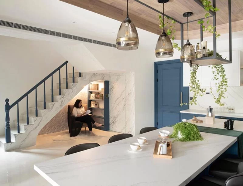 Cette maison moderne dispose d'un petit coin lecture sous l'escalier. Il comprend un mur d'accent en carreaux gris, une étagère bleue personnalisée, un fauteuil confortable, une applique et une paire de prises. #ReadingNook #SeatingNook #UnderStairNook #HomeLibrary