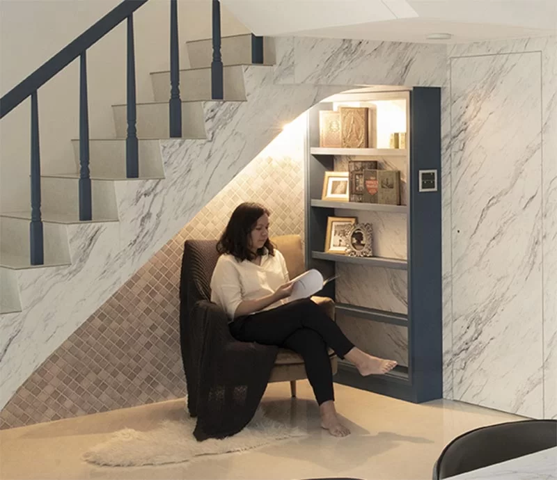 Cette maison moderne dispose d'un petit coin lecture sous l'escalier. Il comprend un mur d'accent en carreaux gris, une étagère bleue personnalisée, un fauteuil confortable, une applique et une paire de prises. #ReadingNook #SeatingNook #UnderStairNook #HomeLibrary