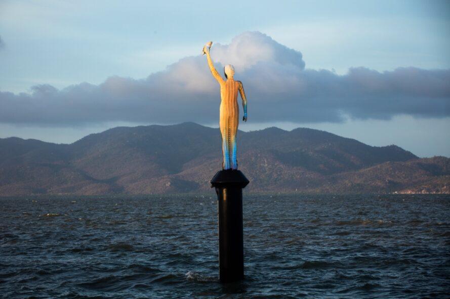 une statue d'une personne avec un bras levé émergeant de l'océan. la statue est jaune et bleue, montrant où la température a changé les couleurs de la statue.