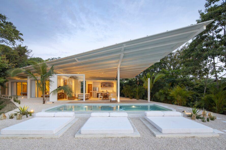 une maison blanche au toit angulaire éclairée de l'intérieur. en face de la maison est une piscine avec trois sièges en forme de lit