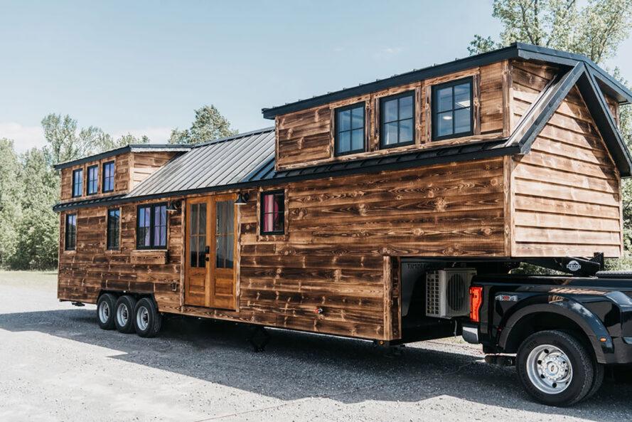 une petite maison sur roues avec un extérieur en bois semblable à une cabine relié par une remorque à un camion