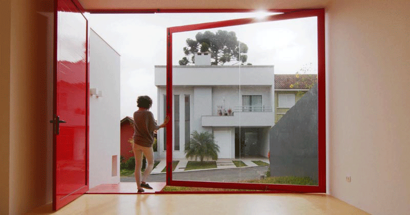 Lorsque la porte d'entrée de cette maison moderne est ouverte, la grande fenêtre pivotante peut être déplacée en poussant le cadre pour l'ouvrir complètement, exposant l'intérieur de la maison au petit patio en herbe. et cour avant. #PivotingWindow #RedFrontDoor #WindowIdeas