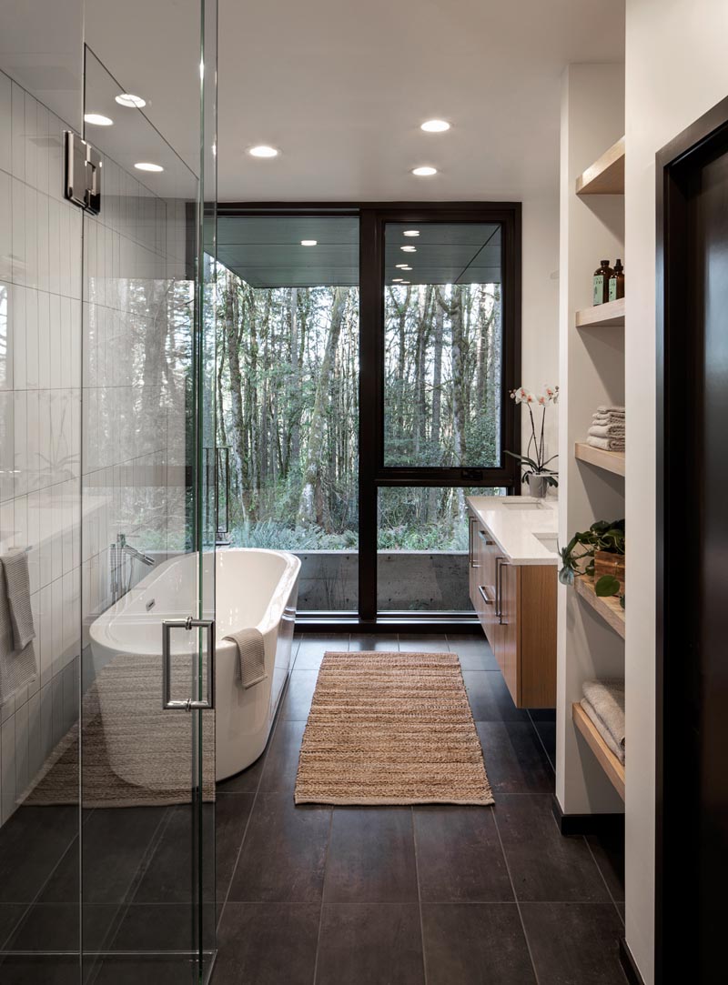 Dans cette salle de bain principale moderne, une baignoire autoportante et la douche sont placées contre un mur, tandis qu'une vanité flottante en bois avec deux lavabos et un mur de rayonnage ouvert sont situés sur le mur opposé. #MasterBathroom #ModernBathroom #BathroomIdeas