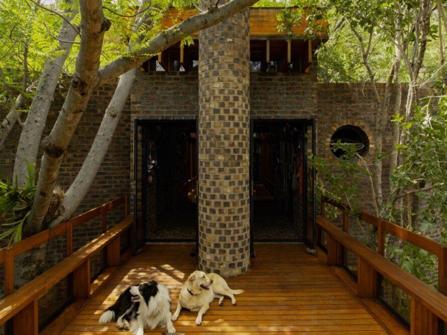 un pont en bois menant à une maison en brique.  au milieu du pont se trouve un pilier de pierre, avec deux chiens allongés devant lui.  des arbres entourent la maison.