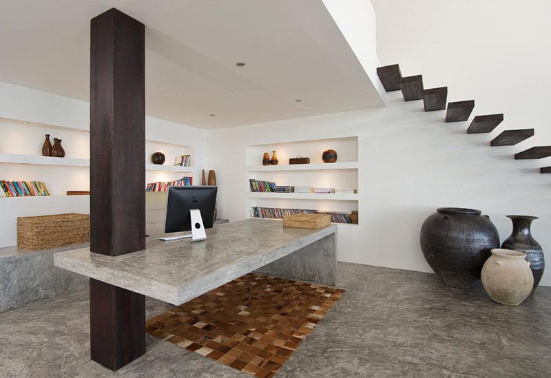 Un bureau en béton utilise une colonne en bois comme support dans ce bureau à domicile qui comprend également des étagères intégrées.