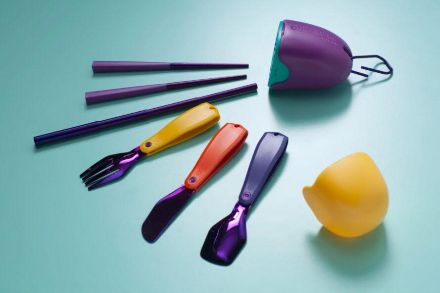 un récipient violet et jaune en forme d'oeuf, un couteau violet et rouge, une fourchette jaune et violette, une cuillère bleue et violette, une paille violette et des baguettes disposées sur un fond bleu.
