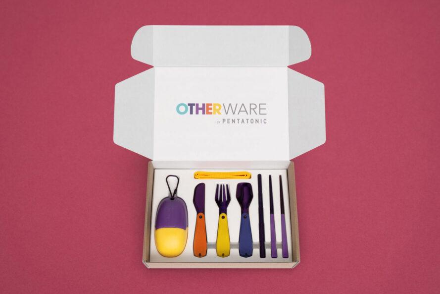 une boîte blanche ouverte sur un fond rose.  le couvercle intérieur supérieur de la boîte indique "AUTRES PRODUITS," et le fond de la boîte contient un récipient violet et jaune en forme d'oeuf, un couteau violet et rouge, une fourchette jaune et violette, une cuillère bleue et violette, de la paille violette et des baguettes.