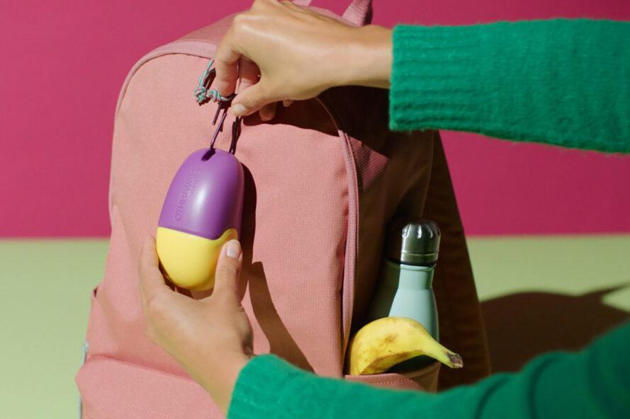 une paire de bras dans un pull vert attachant un récipient violet et jaune en forme d'oeuf à un sac à dos rose